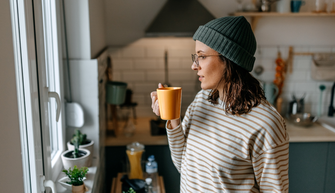 woman with coffee cup looking through window while standing in kitchen at home