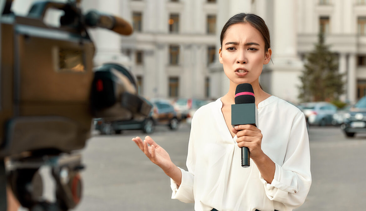 A news reporter holds an external microphone while being filmed for a news segment.