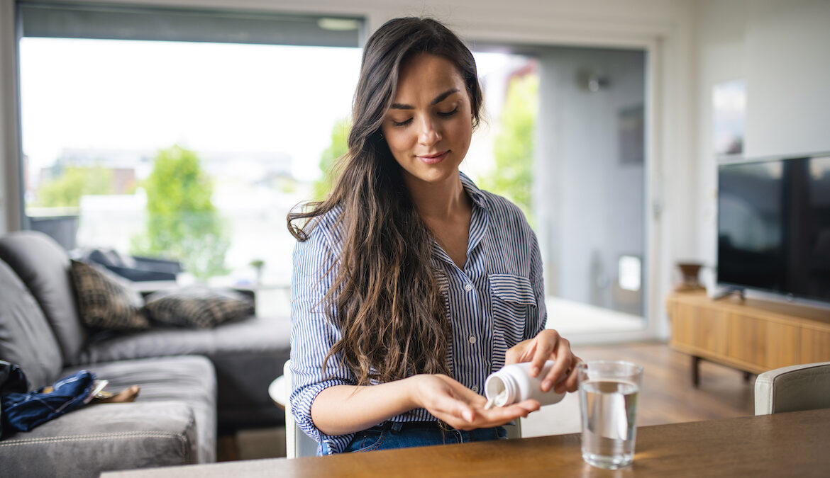 A beautiful brunette woman pours out white supplement pills into her hands while standing in her kitchen.