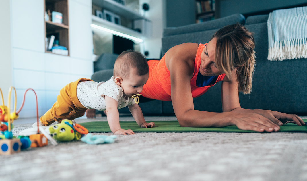 Woman doing forearm plank while baby plays next to her at home.