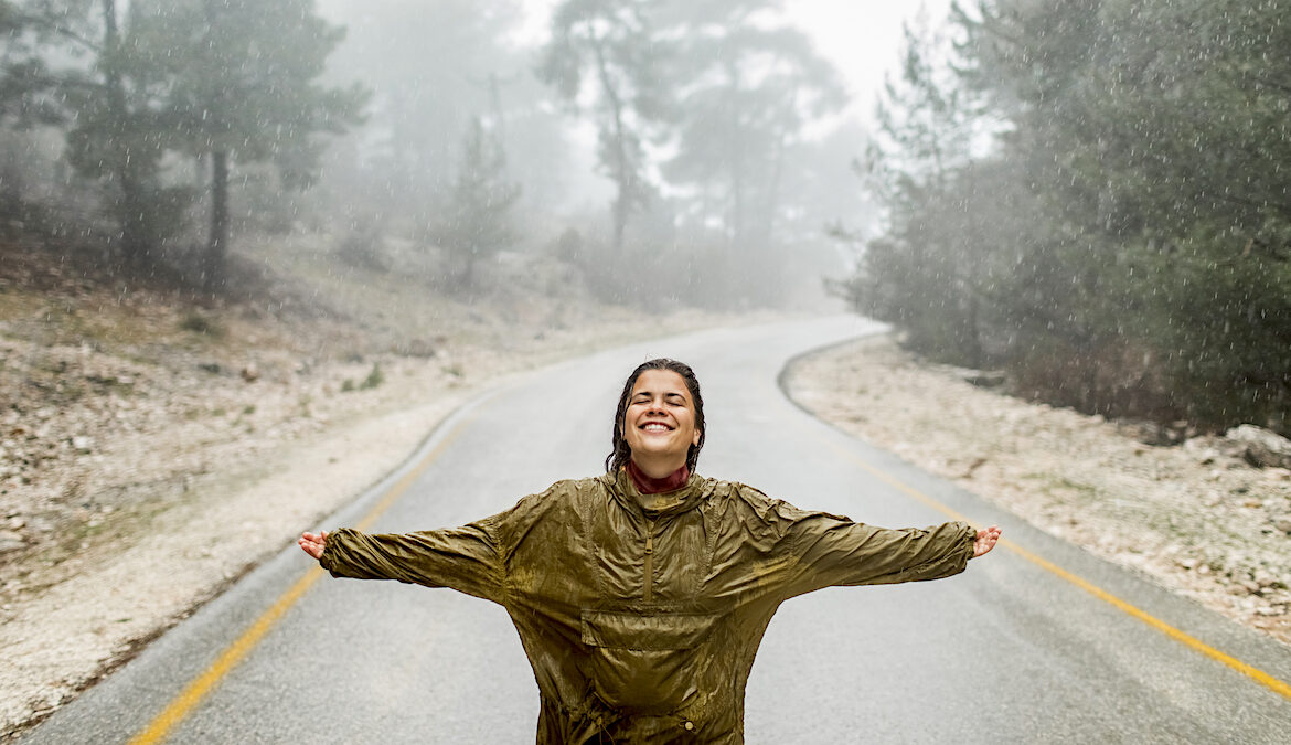 A smiling woman stands in the middle of an empty road with her hands outstretched as rain pours down.