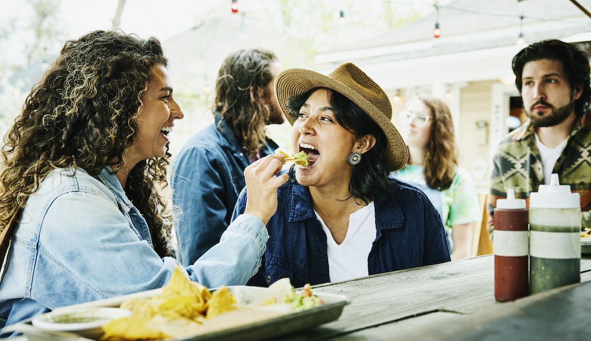 A laughing woman feeds her friend a chip with guacamole while eating at a food truck.