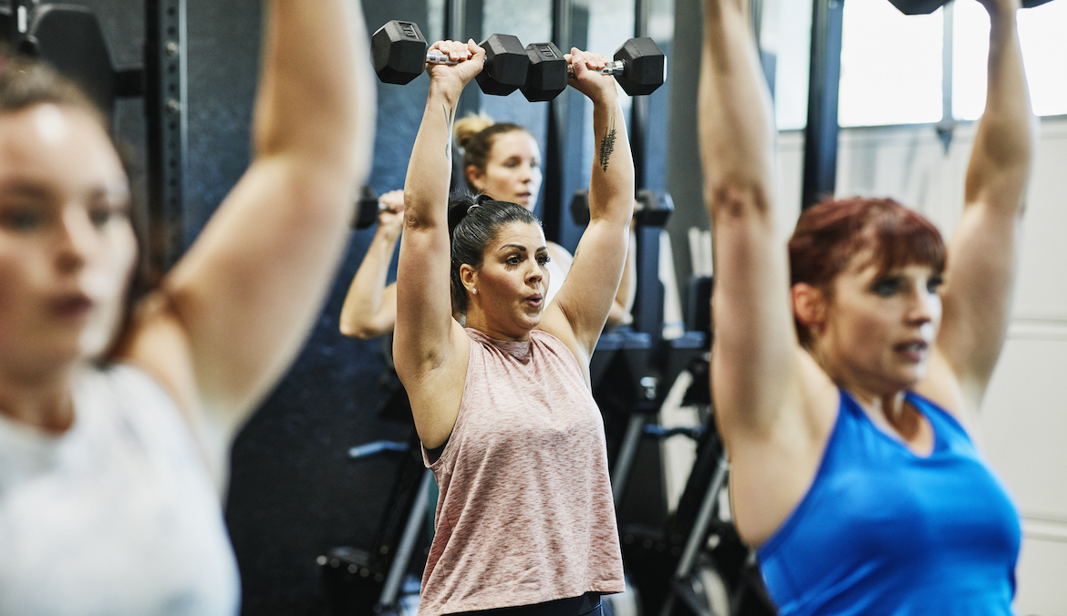 Women's fitness class doing overhead dumbbell presses in gym