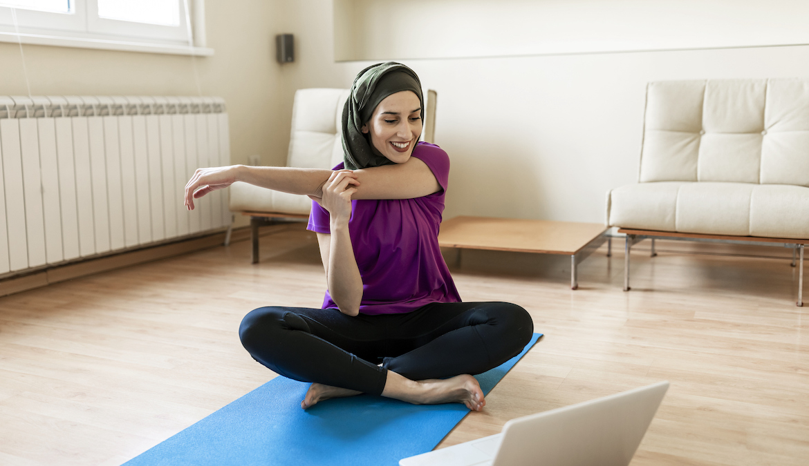 A Muslim woman is looking at a video lesson of yoga on her laptop and exercising at home.