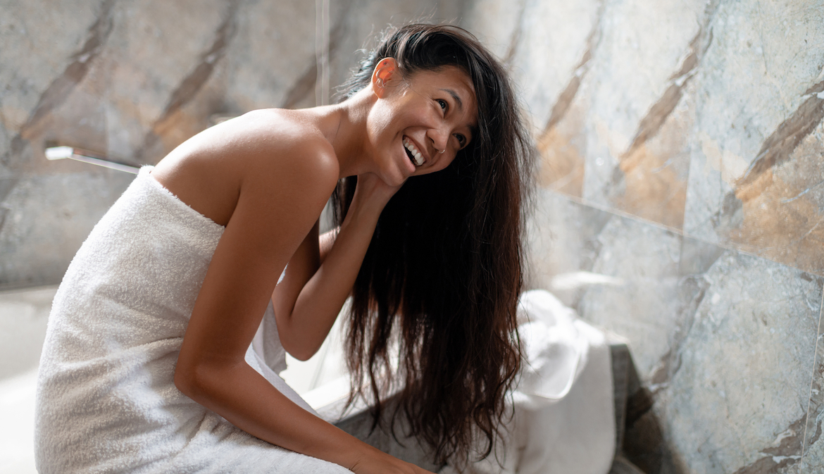 Happy young woman genuinely laughing while detangling her hair on the edge of the bath tub