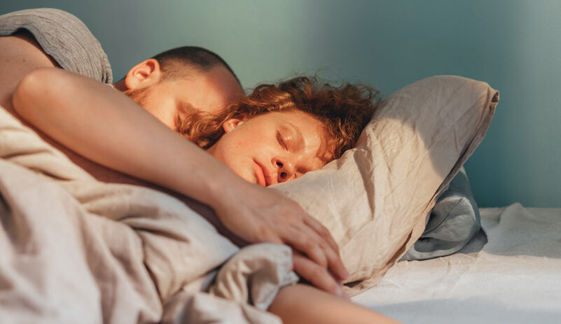 A young woman sleeps while embraced by a man in bed.