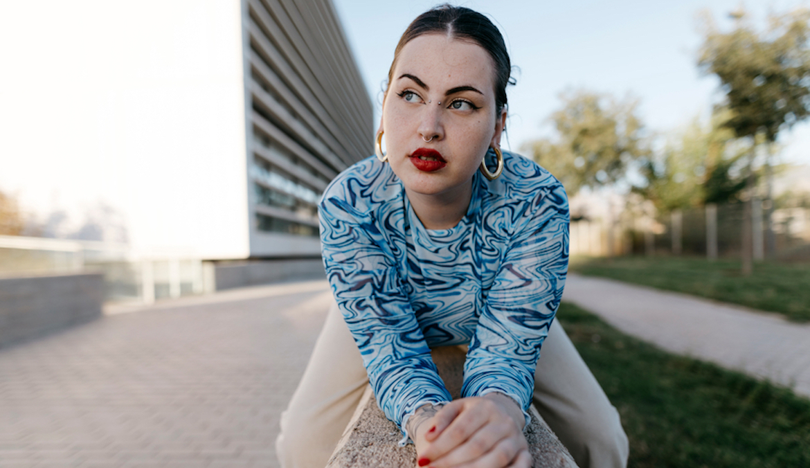 thoughtful woman in stylish clothes looking away while sitting and leaning forward on concrete border in city on summer day