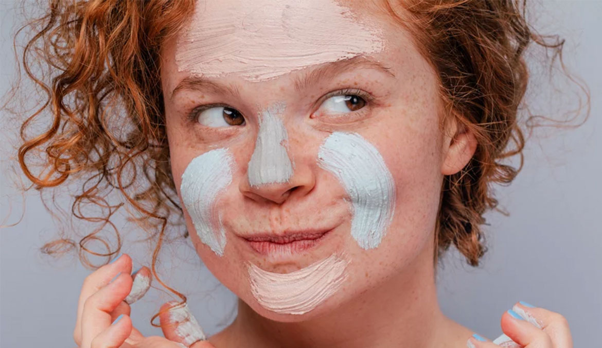 A portrait of a woman with red hair using a clay mask on her face.