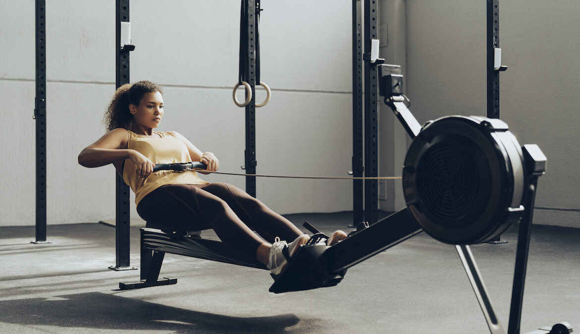 A young woman leans back while using a rowing machine inside of a gym, wearing a pair of rowing shoes