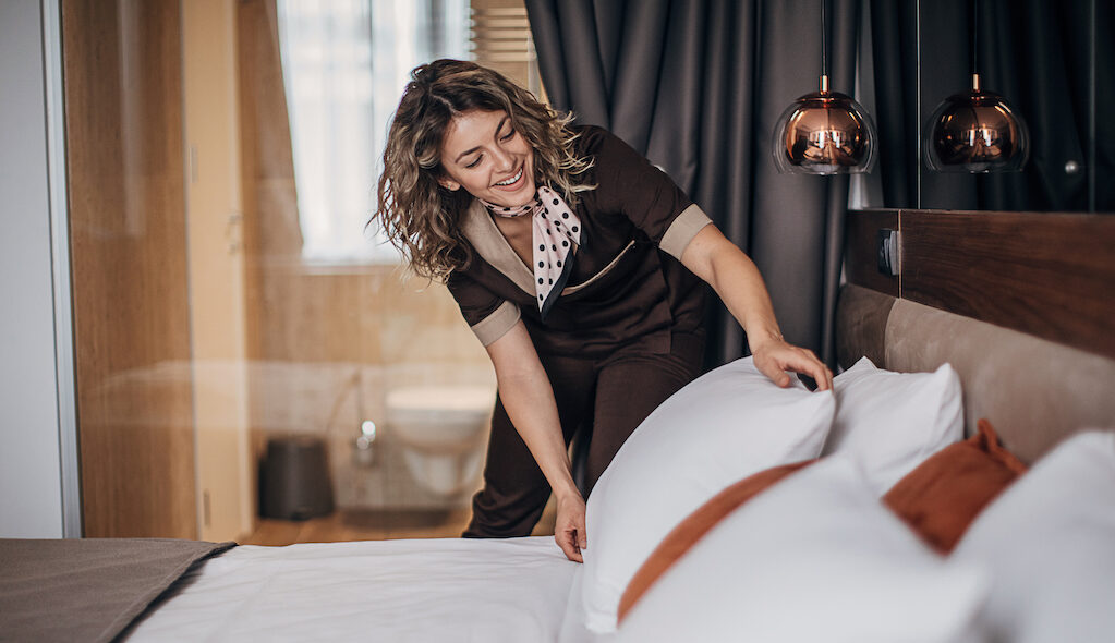 A pretty female hotel housekeeper fluffs a hotel pillow while smiling.