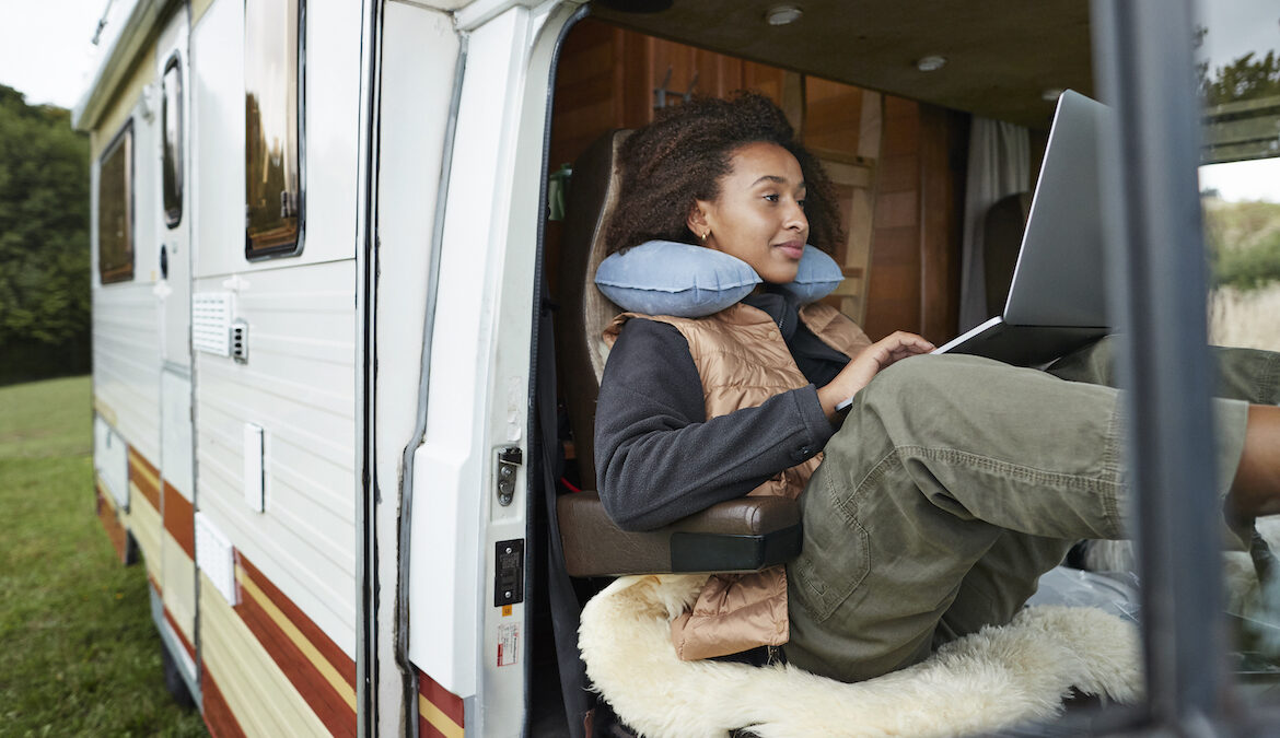 A beautiful young woman sits in her van while typing on a laptop and wearing an airplane pillow around her neck.