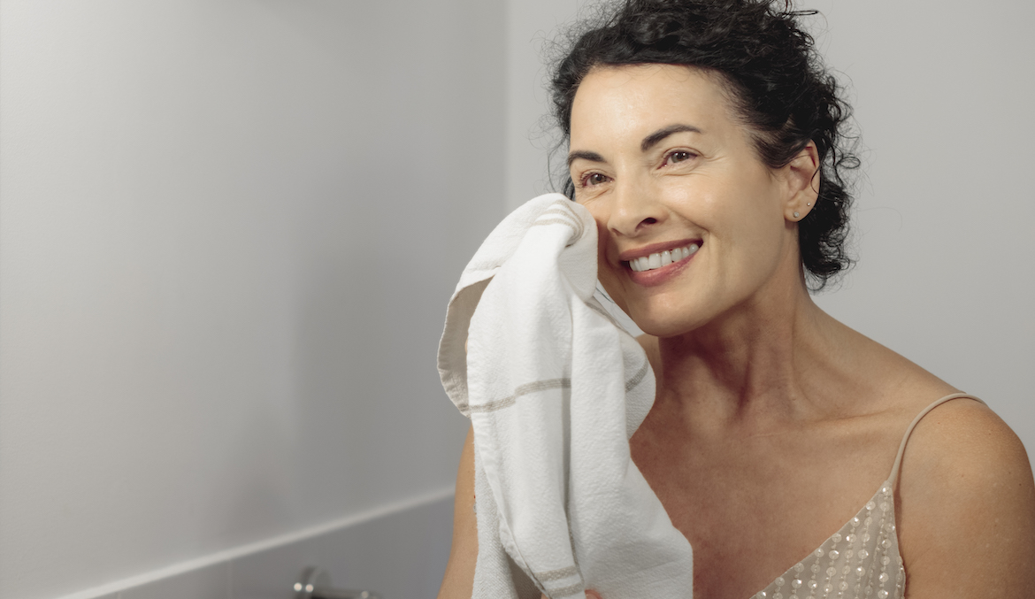 Woman in her 40s with brown, curly hair smiling while wiping her face with a towel in her bathroom.