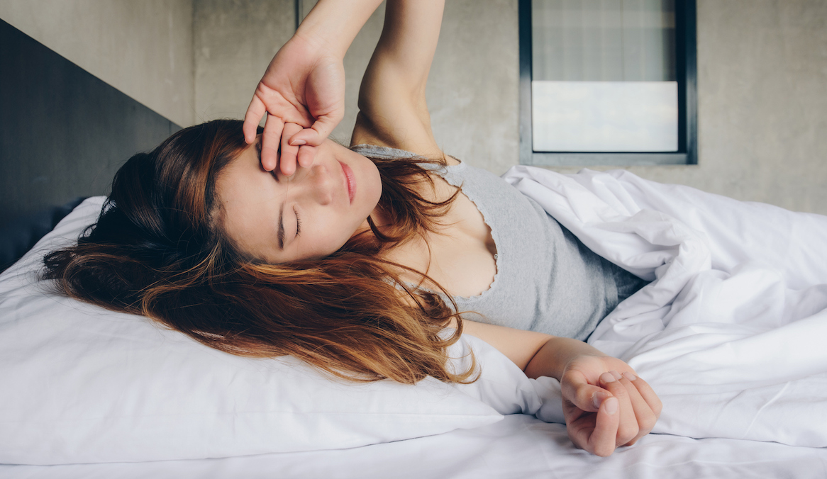A woman in a gray tank top lies in a bed and is waking up.