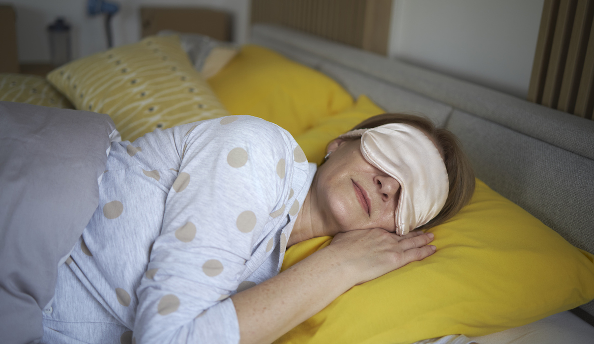 A woman in polka dot pajamas sleeps on a bed with yellow sheets and wears a satin eye mask over her eyes.