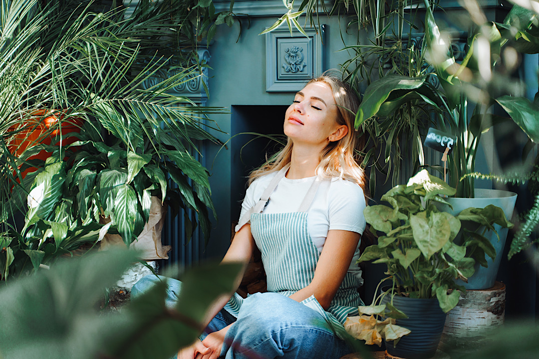 Woman sitting with eyes closed surrounded by plants.