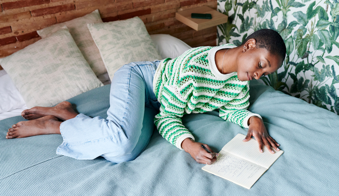 young black woman writing in her journal while spending a relaxing afternoon lying on her bed at home