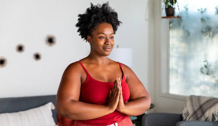 A beautiful plus size black woman with natural hair texture stands smiling in an energy cleanse stance with her prayer hands over her chest.