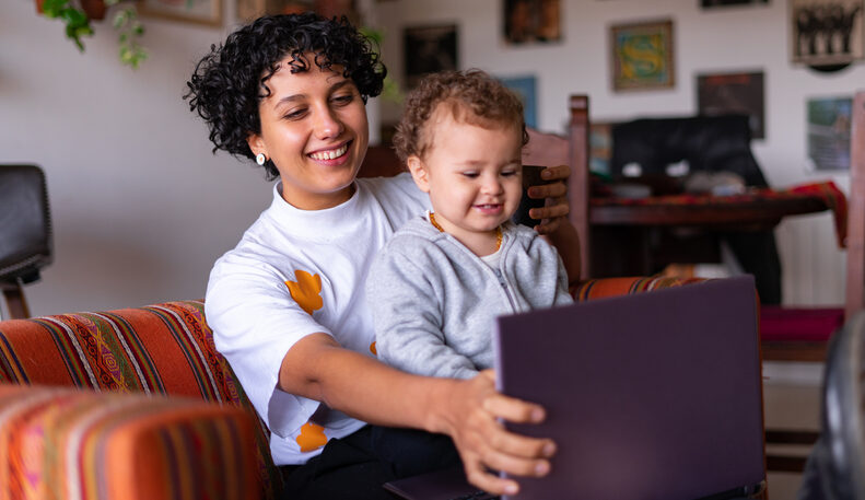 A young woman smiles while holding her baby on her lap and adjusting the screen of her laptop.