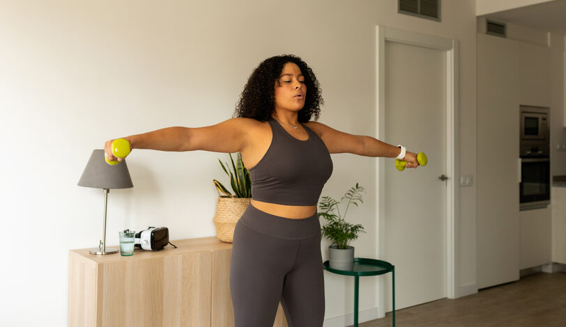 A curvy woman wearing gray athletic clothing breathes deeply while lifting dumbbells in a side lateral raise.