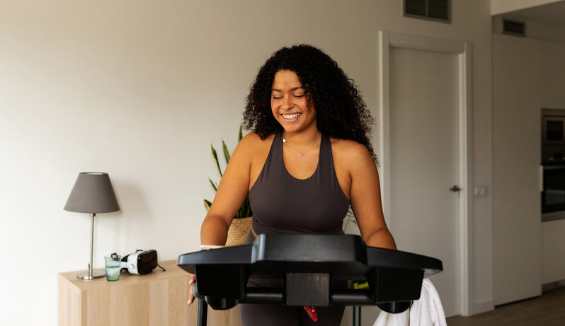 A smiling young woman wearing athletic clothing walks on a smart treadmill inside of her home.