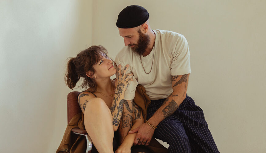 A young, beautiful tattooed couple smiles at each other and holds each other while having matching couples tattoos.