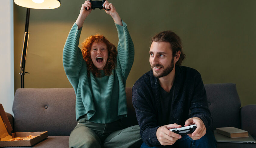 A young optimist woman and her pessimist boyfriend laugh and sit on a couch while playing video games.