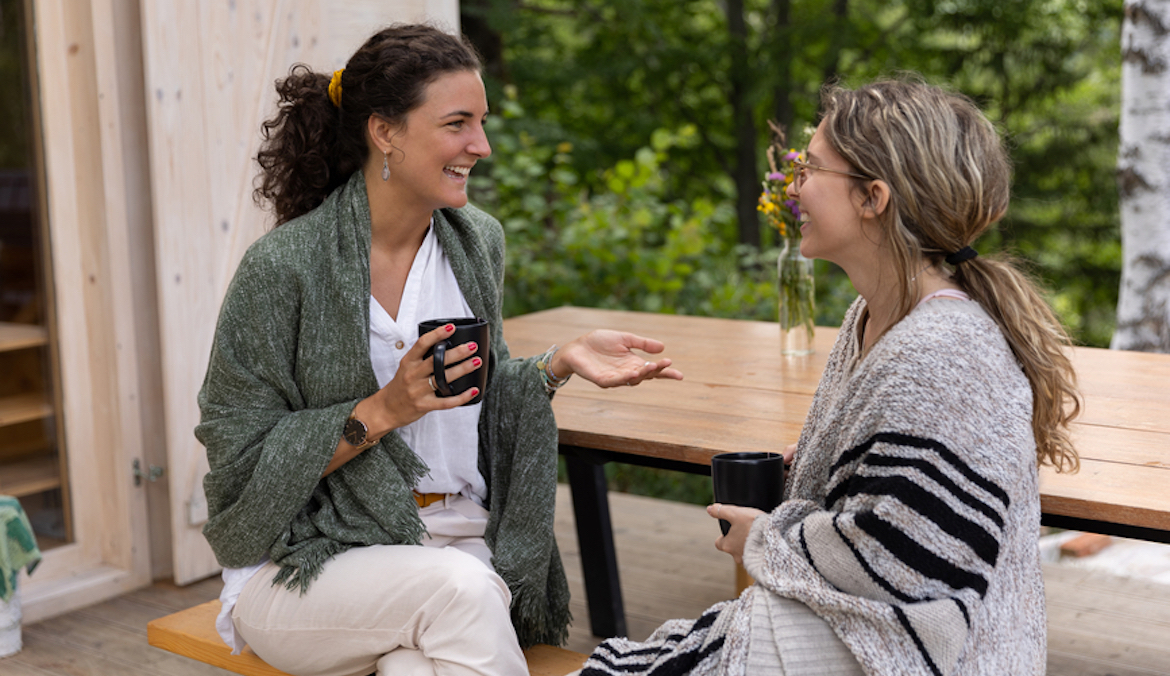 two female friends sitting on a porch of a cottage house, spending their time chatting and drinking coffee