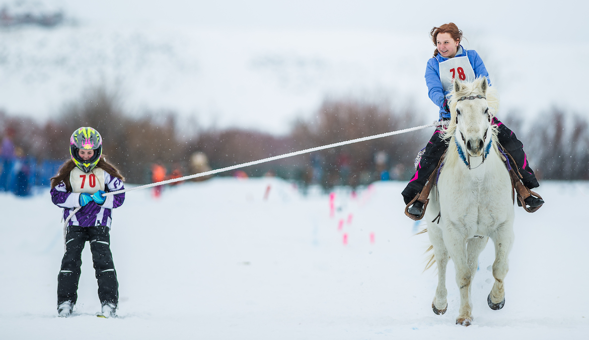 A woman skiing while holding a rope connected to a horse, being ridden by another woman.