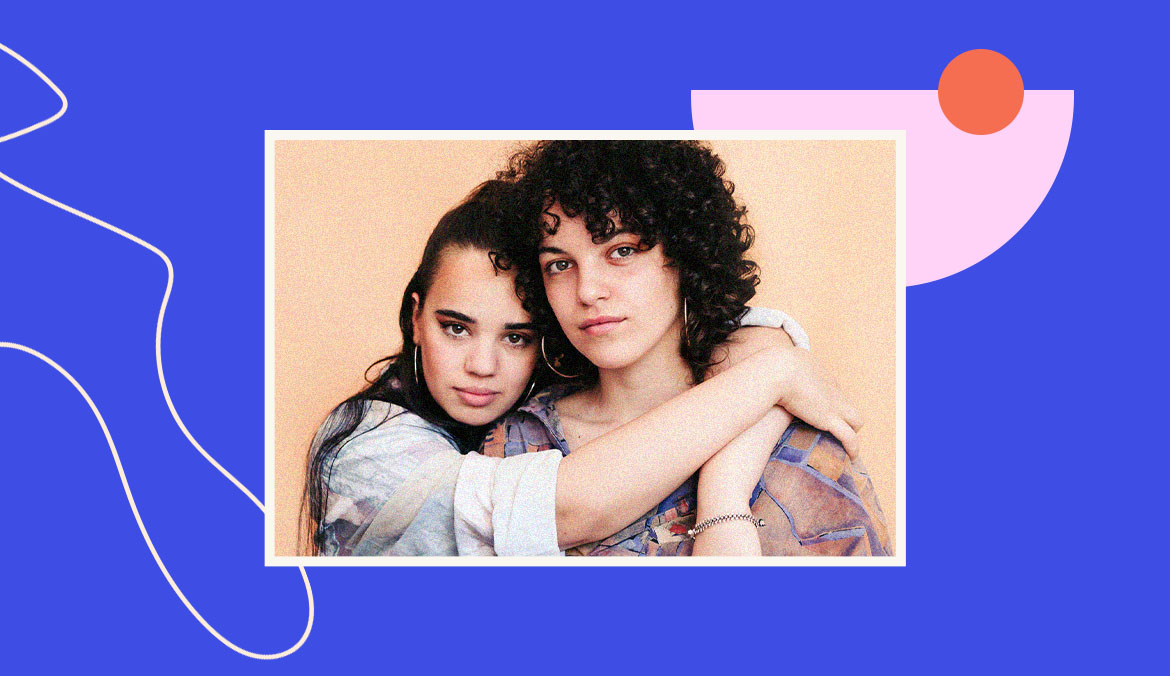 Two young women lean on each other in an embrace against a graphic design background.