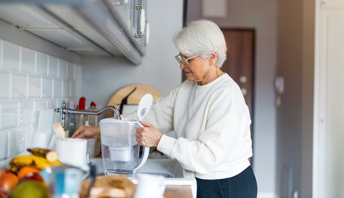 A senior caucasian woman fills up a water filter while standing in a white kitchen.