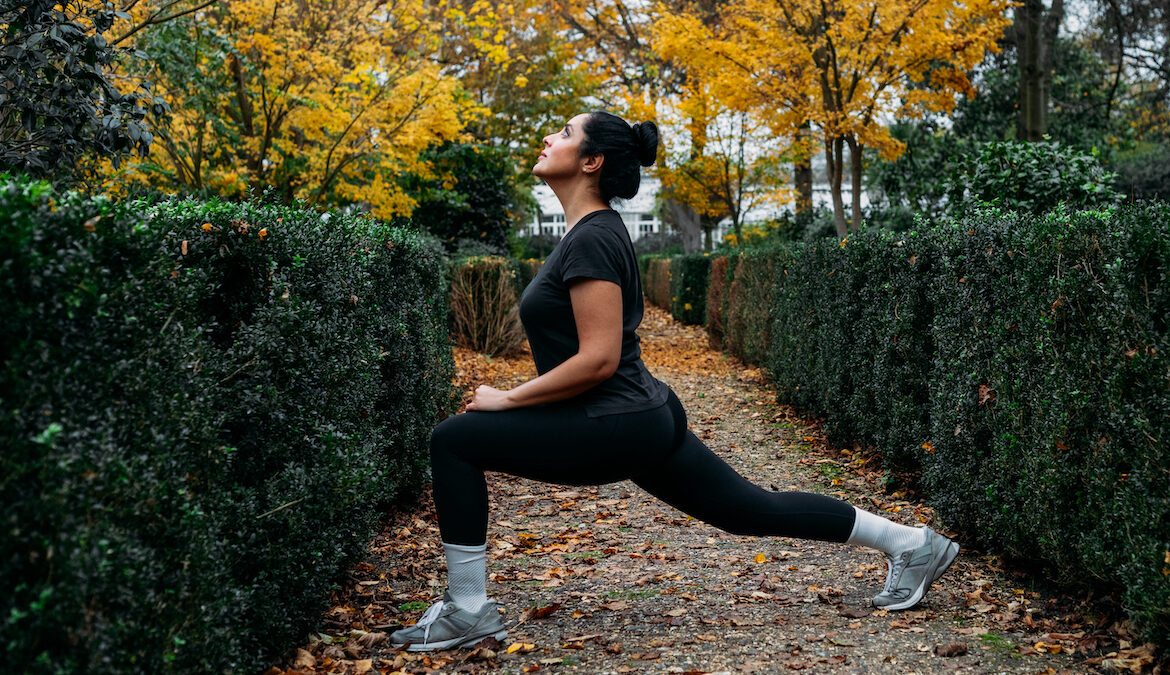 A young athletic woman stretches her hip flexors in a deep lunge while outside on a walking trail.