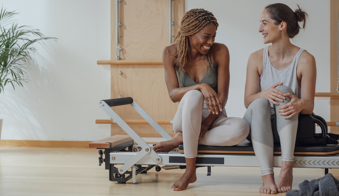 Two beautiful smiling women in sportswear sitting on pilates reformer.