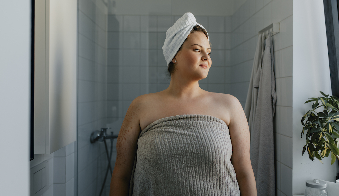 A smiling woman stands in the bathroom with a towel.