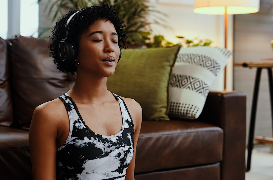 Young Black woman sitting on floor in living room wearing headphones and practicing breathing exercises.
