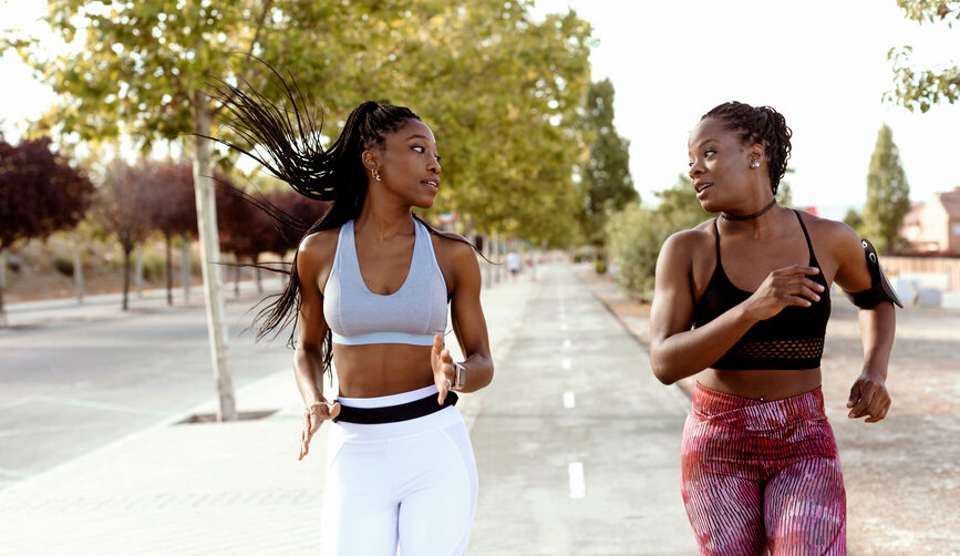 Two young black women run alongside each other outdoors on a street, racing each other, symbolizing competition and envy.