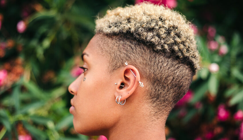 Side profile photo of a stylish person with shaved hair and several ear piercings.