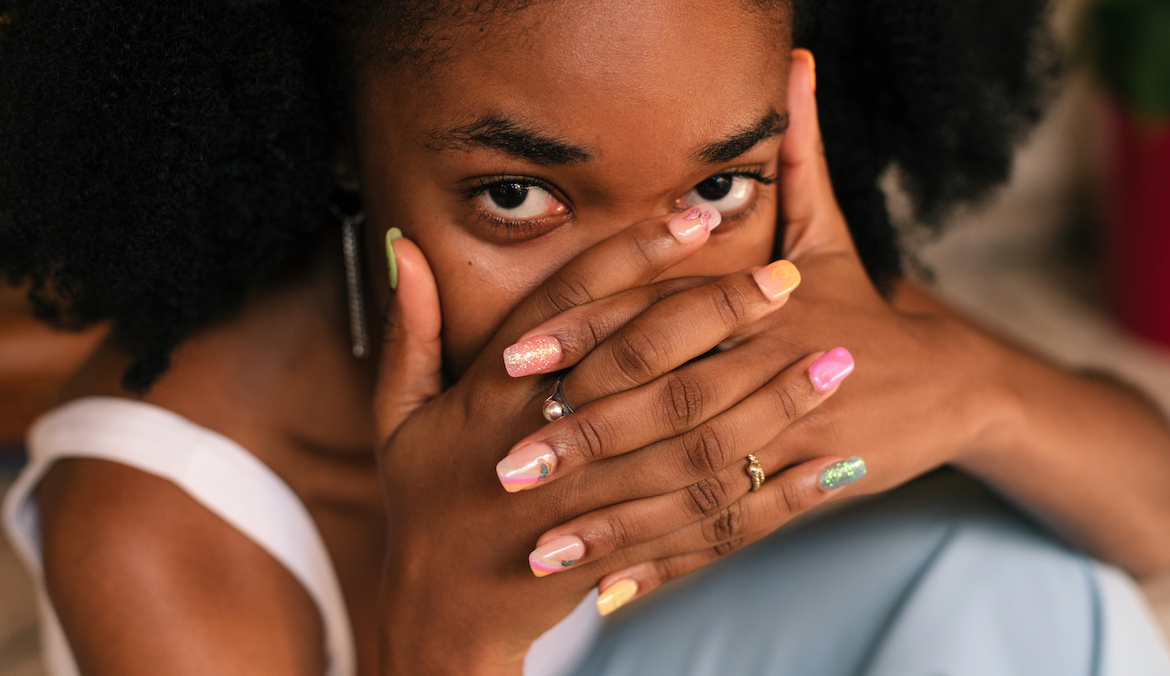 Young black woman with her hands covering her face, with a colorful and trendy manicure, looking at camera.