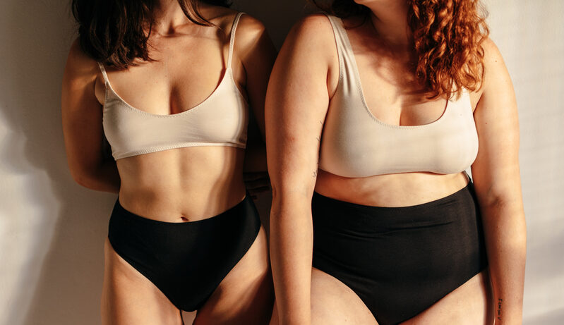 Two women of different body sizes stand side by side wearing neutral bras and underwear.