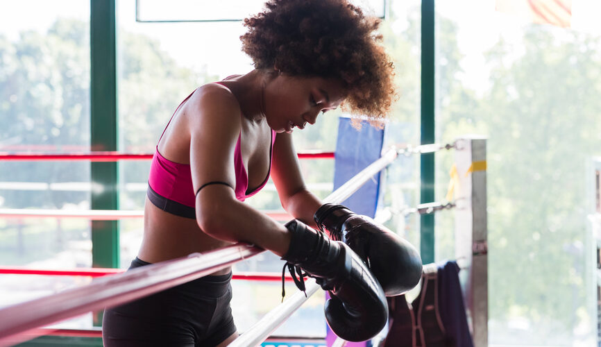A strong young black boxer rests her arms over the side of a boxing ring, tired from her workout.