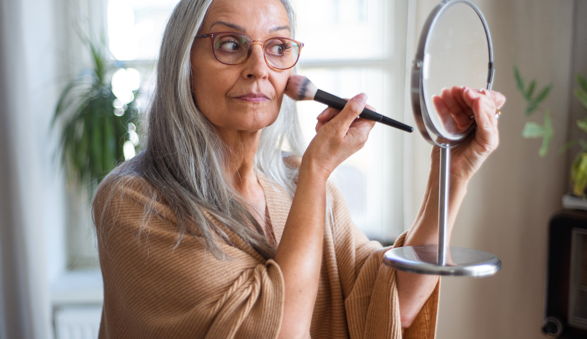woman with grey hair and glasses applying the merit beauty flush balm in the mirror