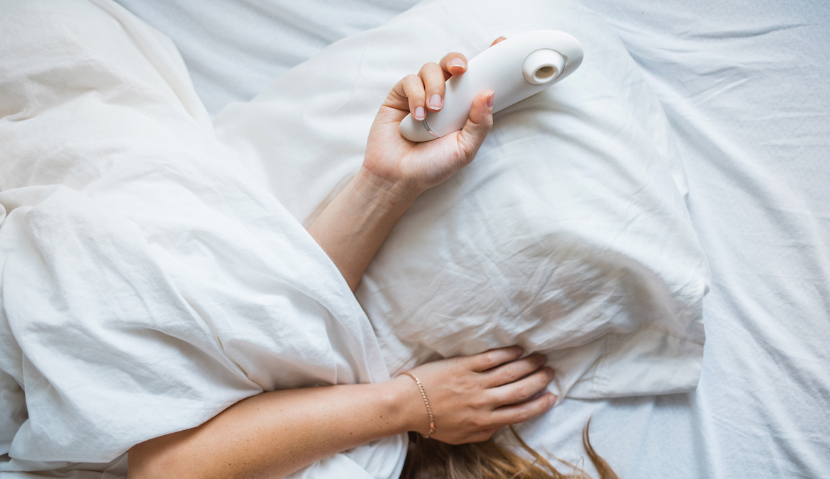 A woman holding a vibrator while under the covers in a bed with white sheets.