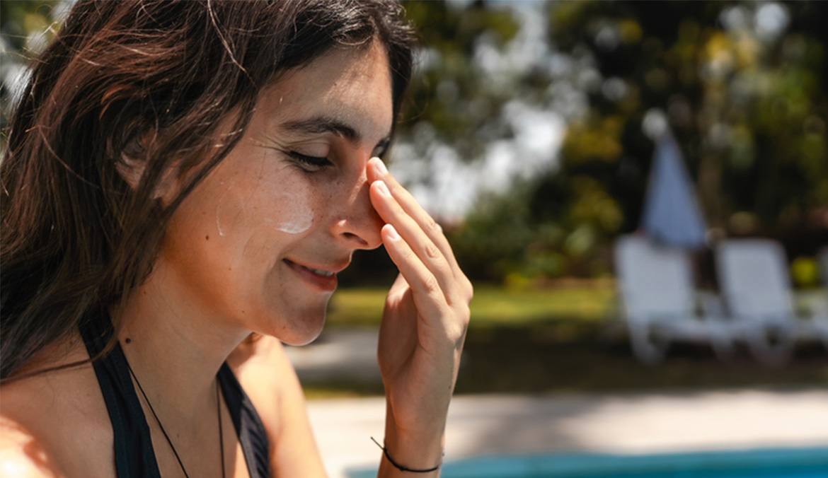 A woman sitting by a pool applying sunscreen to her face.