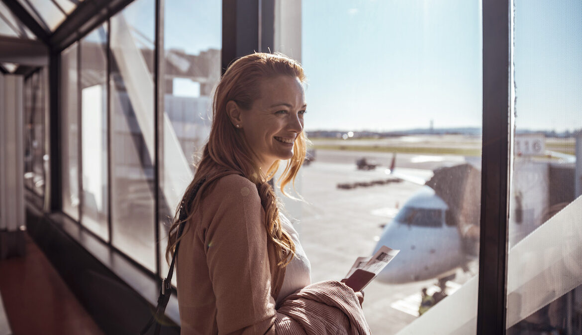 A red-haired young woman smiles and looks at planes from a large window in an airport.