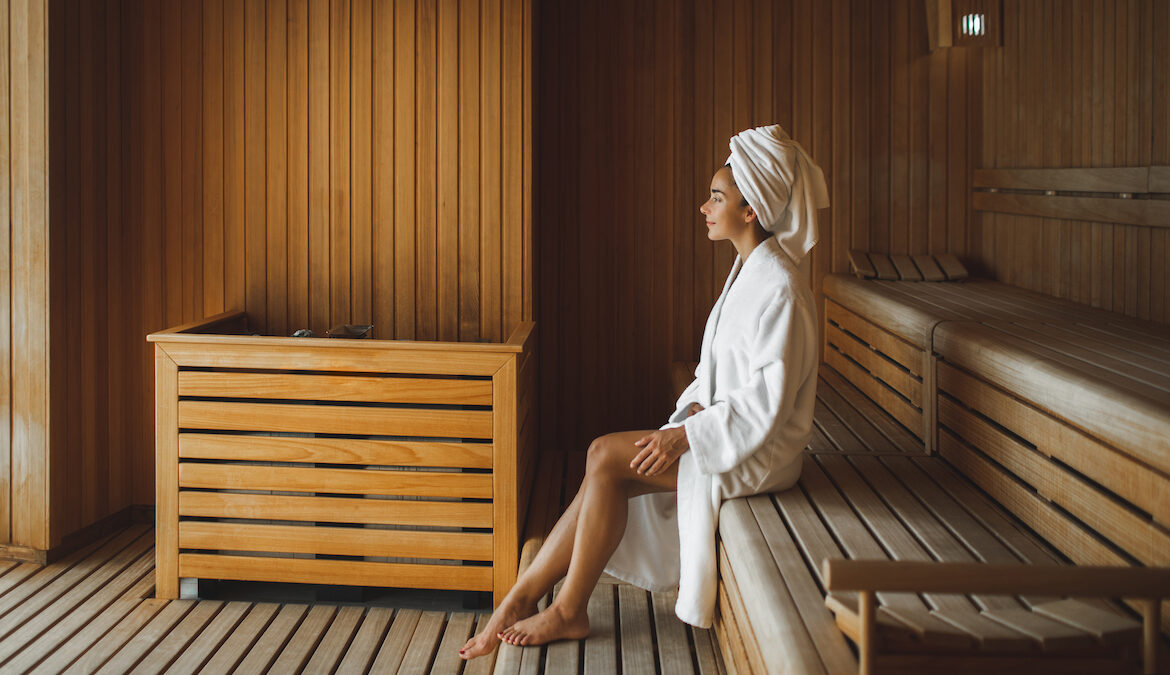A young woman wearing a white bathrobe and white towel on her head sits in a hot sauna.