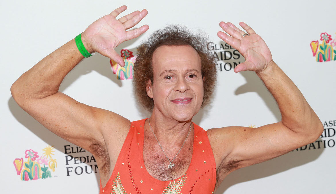 Fitness icon Richard Simmons stands in front of a backdrop while smiling for a press photo.