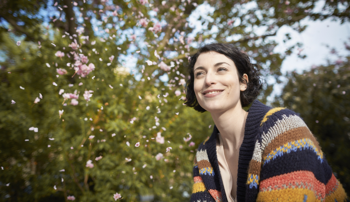 Woman in her mid-20s with short hair wearing a colorful striped sweater smiling in front of a blossoming spring tree.