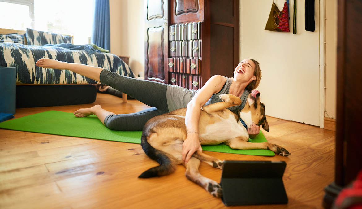Young woman petting her dog and laughing while trying to do an online workout at home