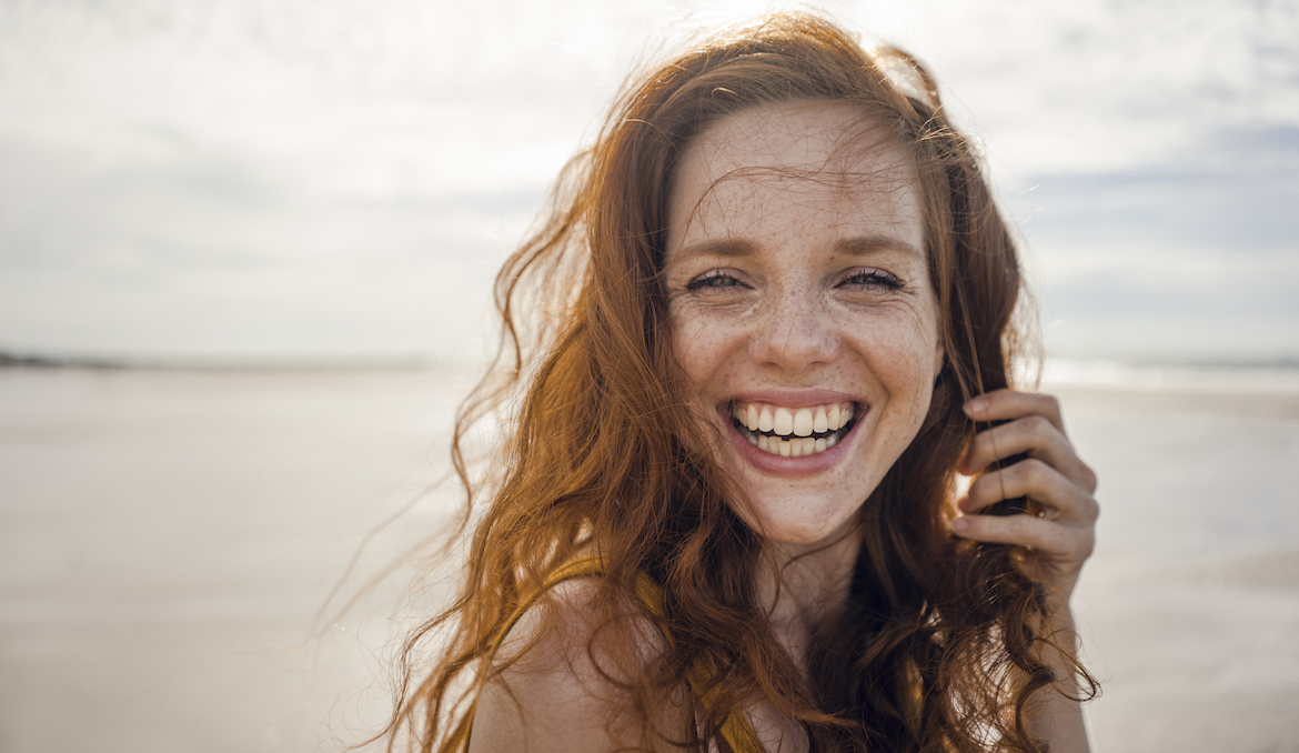 A young redheaded woman smiles and looks into the camera on the beach.