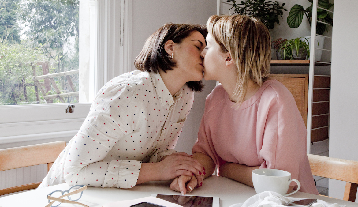 Two women sit at a table and kiss.