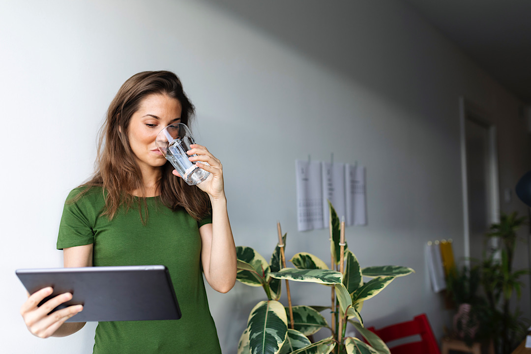 woman drinking water while using digital tablet against wall at home